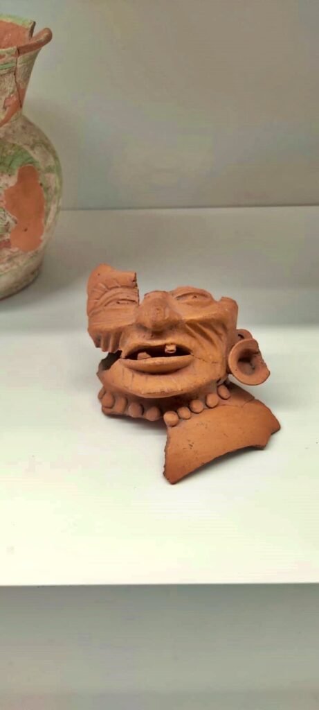 Fragmento de figurilla de barro con rostro humano y collar, sobre repisa blanca; al fondo se ve parte de una vasija.