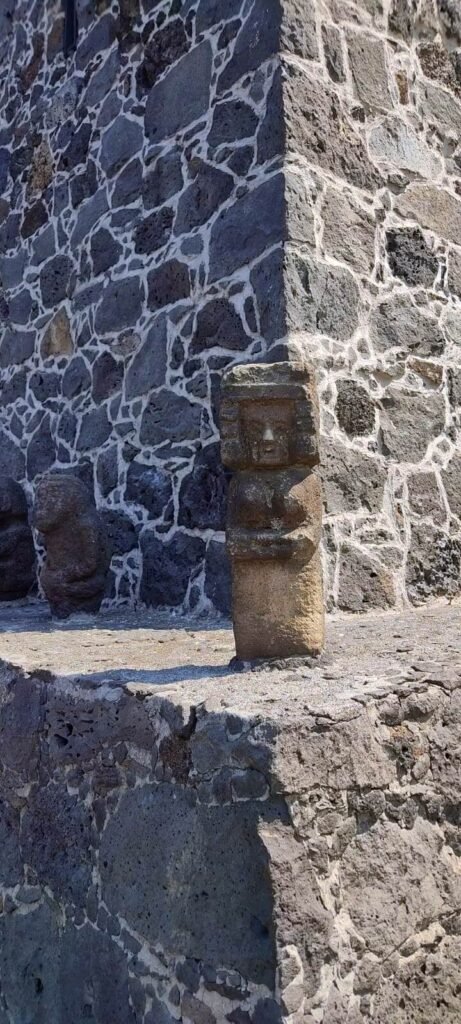 Pequeña escultura de piedra en primer plano sobre un escalón, junto a muro de piedra volcánica del museo, con otra figura al fondo.