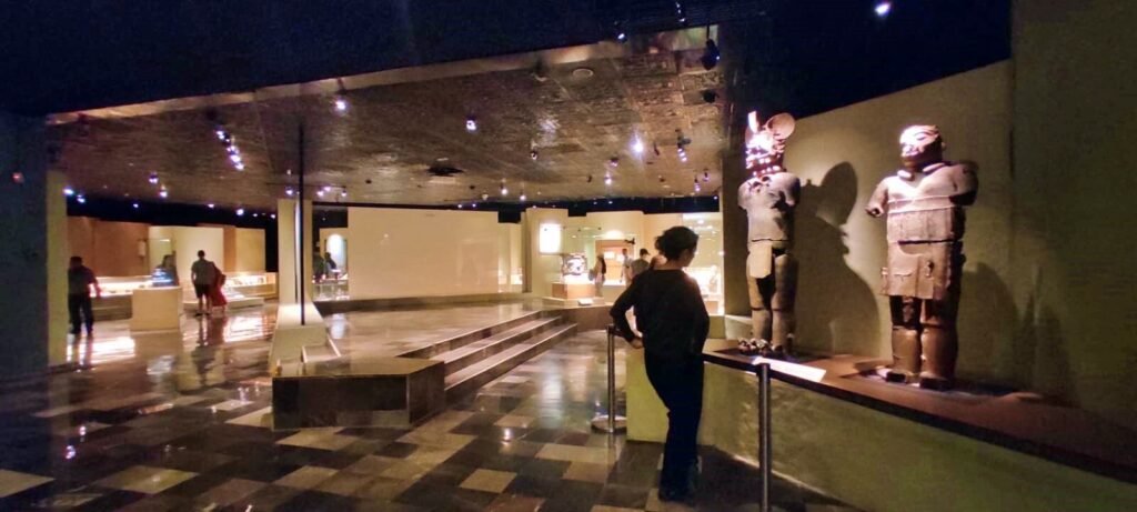 Sala del Museo del Templo Mayor con vitrinas iluminadas, esculturas y visitantes recorriendo el espacio.