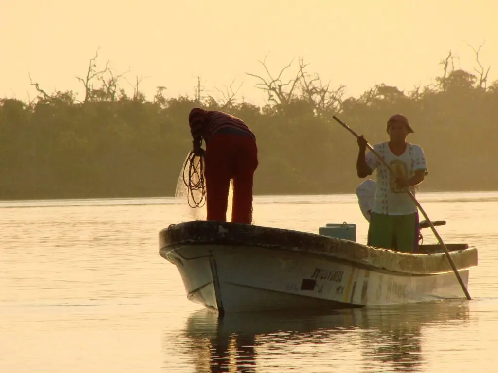 Pescadores en lancha al amanecer, uno lanza red y otro rema en agua calma.