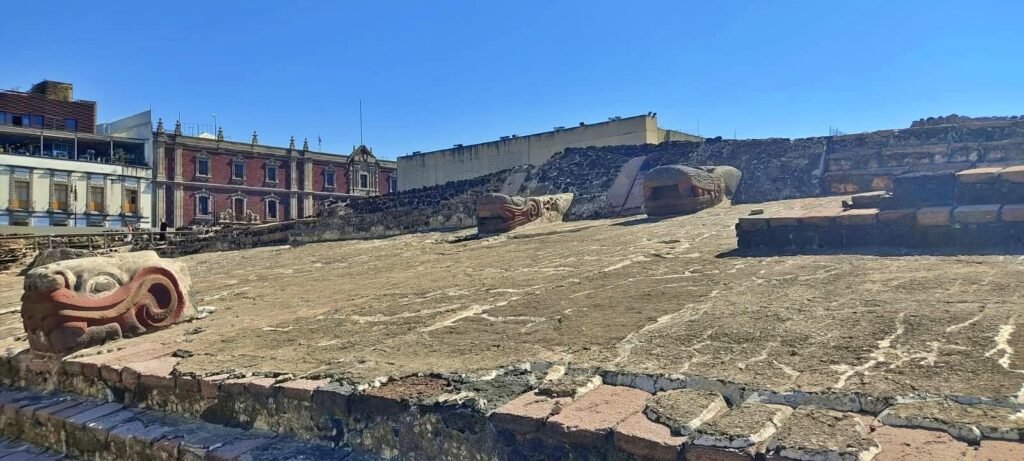 Talud y plataformas del Templo Mayor con cabezas de serpiente decoradas, con edificios del Centro Histórico alrededor, CDMX.