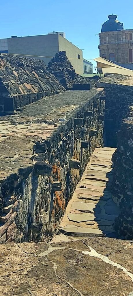 Pasillo estrecho entre muros de piedra del Templo Mayor, con vista hacia edificios del Centro Histórico, CDMX.