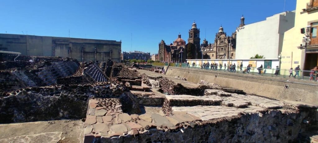 Recorrido por las ruinas del Templo Mayor con la Catedral Metropolitana al fondo y visitantes en la pasarela, CDMX.