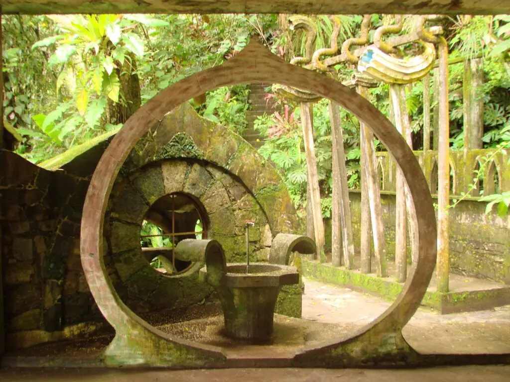 Arco circular de concreto con fuente central y pasillos simétricos cubiertos de musgo en el Jardín Surrealista.